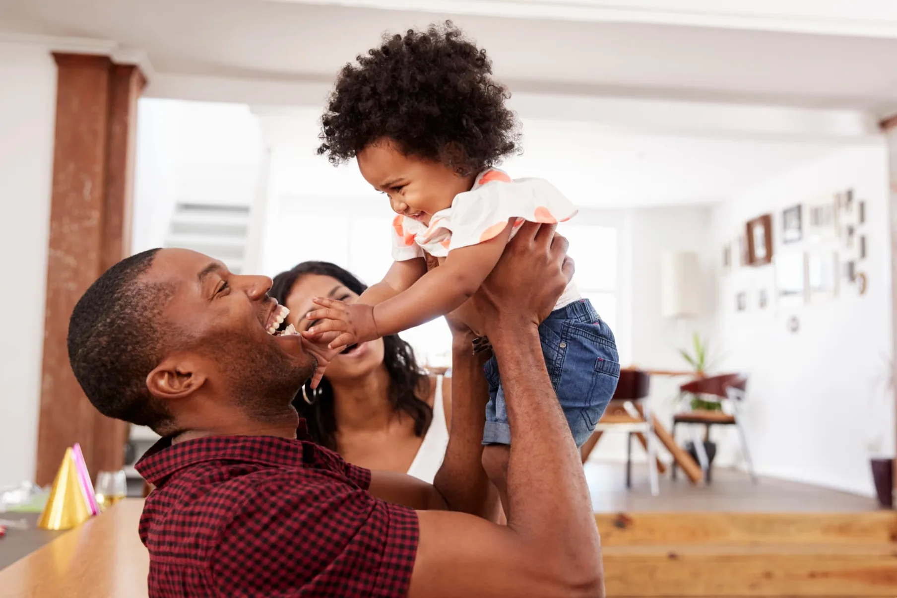 A father lifting his laughing toddler in the air at home while her mother looks on.