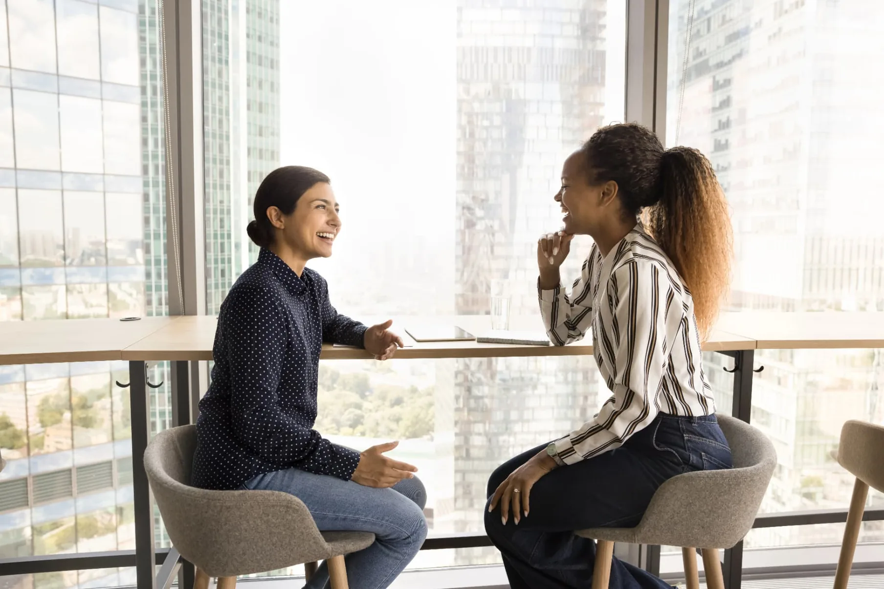 Two colleagues talking across a high table by a window in a city office.
