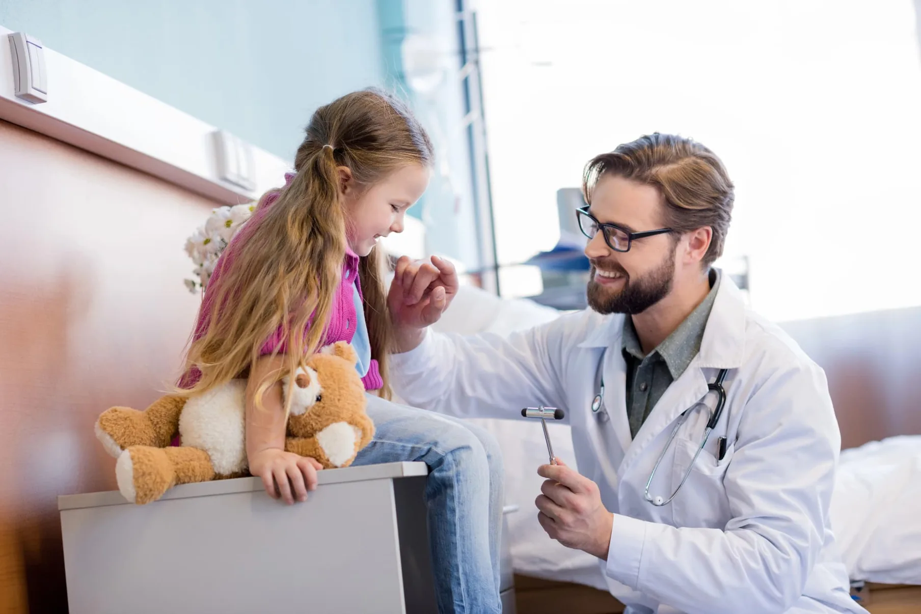 A doctor kneeling beside a young girl and her teddy bear during a check-up.