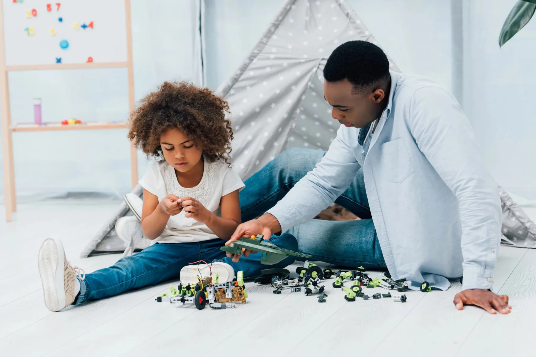 A father and his young daughter building a toy robot together on the floor at home.