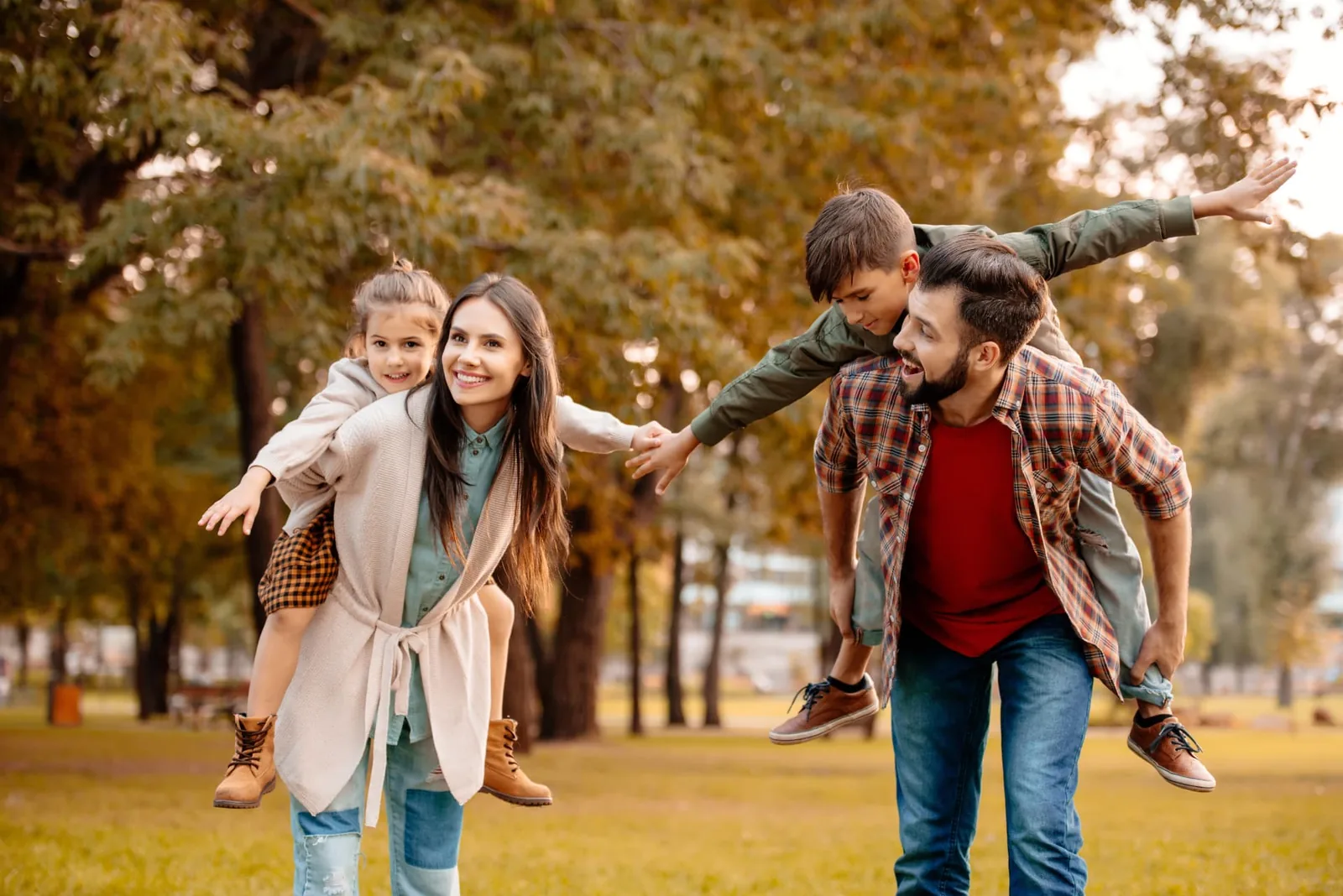 Two parents giving their young son and daughter piggyback rides through a park in autumn.